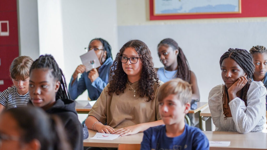 Leerlinge die rustig en aandachtig voor zich uit kijkt in de klas, terwijl medeleerlingen rond haar in beweging zijn